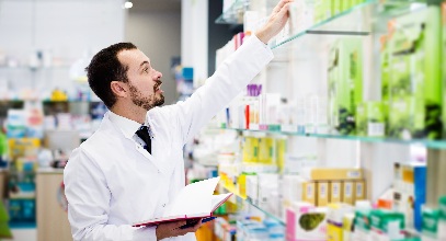 dark-haired male pharmacist in a white coat reaching for something on a high shelf of a pharmacy referencing a notebook in his other hand