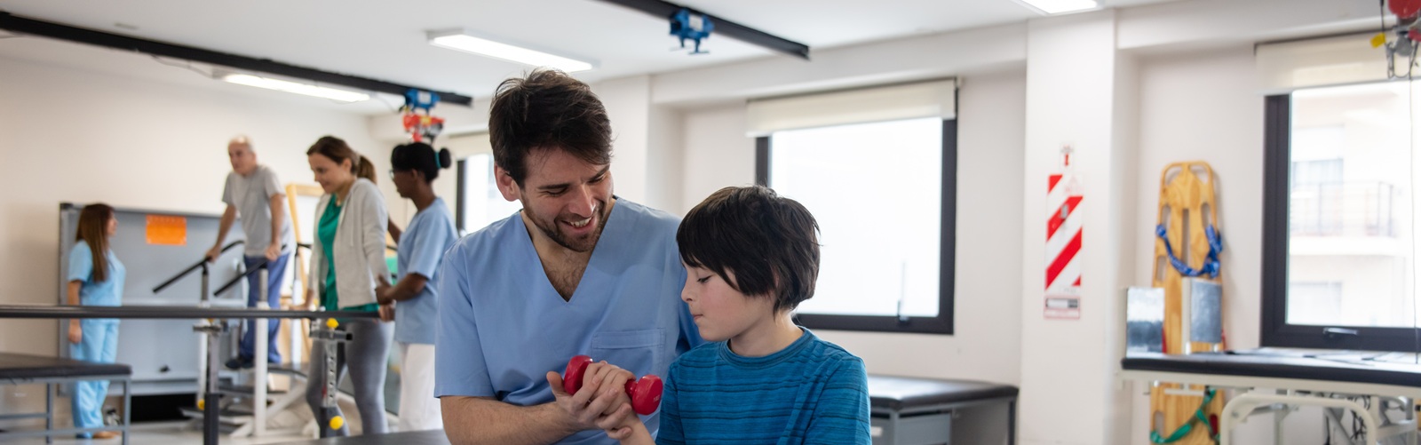 man in blue scrubs helping a small male child in a teal tshirt hold a weight while they are in a medical rehabilitation clinic