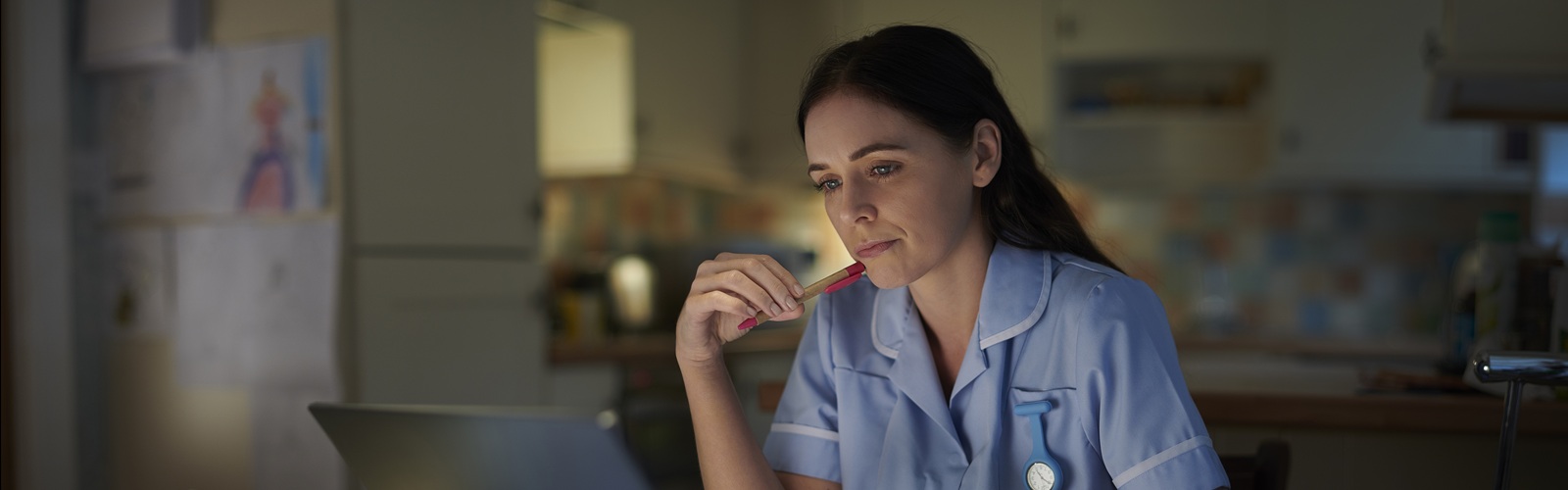 healthcare worker in blue scrubs and long brown hair looking at her laptop surrounded by open books