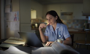 healthcare worker in blue scrubs and long brown hair looking at her laptop surrounded by open books