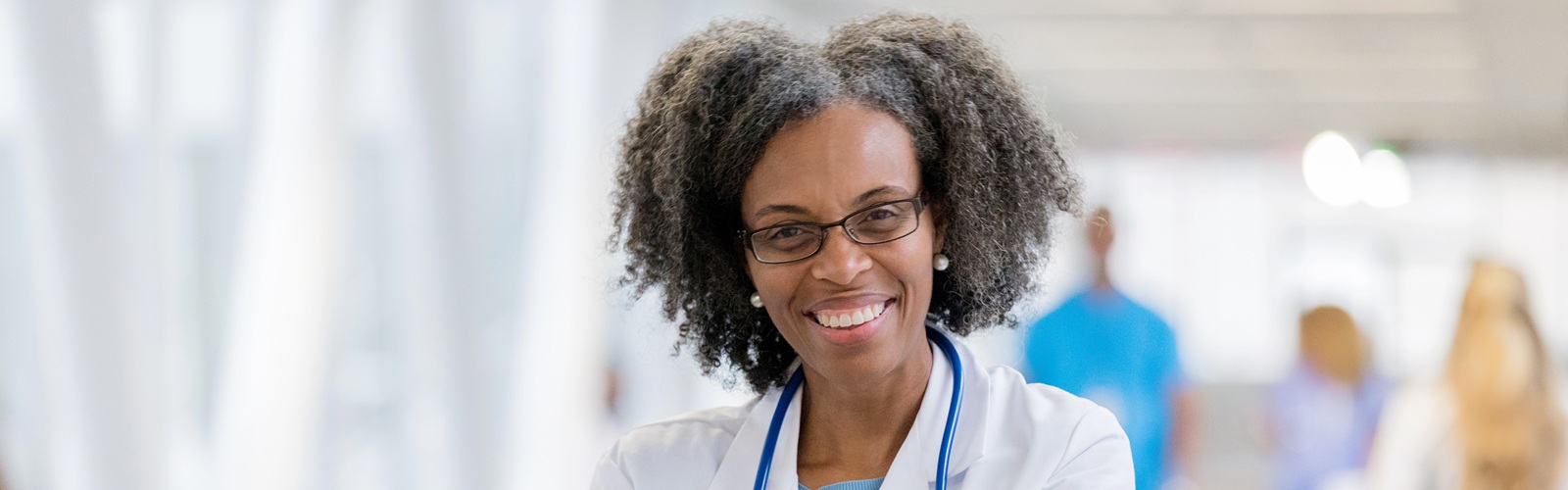 african american nurse in a white lab coat and stethoscope standing in a hospital hallway with her arms crossed smiling