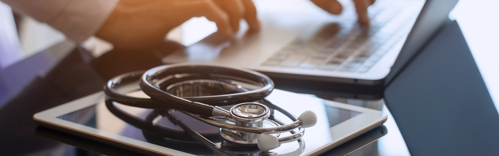person typing on a laptop with a tablet and a stethoscope sitting next to them on the glass table