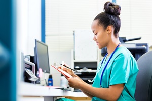 young woman in scrubs sitting at the computer looking at notes on a clipboard