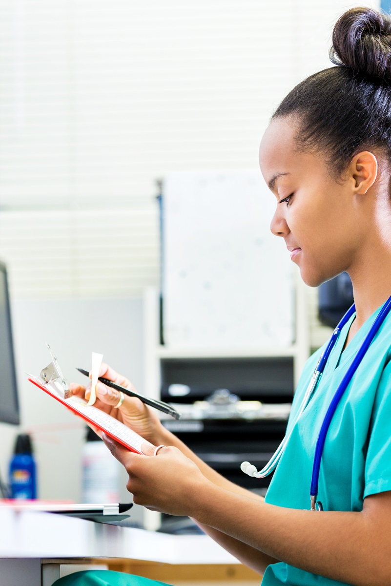 young woman in scrubs sitting at the computer looking at notes on a clipboard