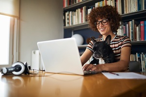 woman with brown curly hair and glasses smiling sitting at her laptop with a french bulldog on her lap