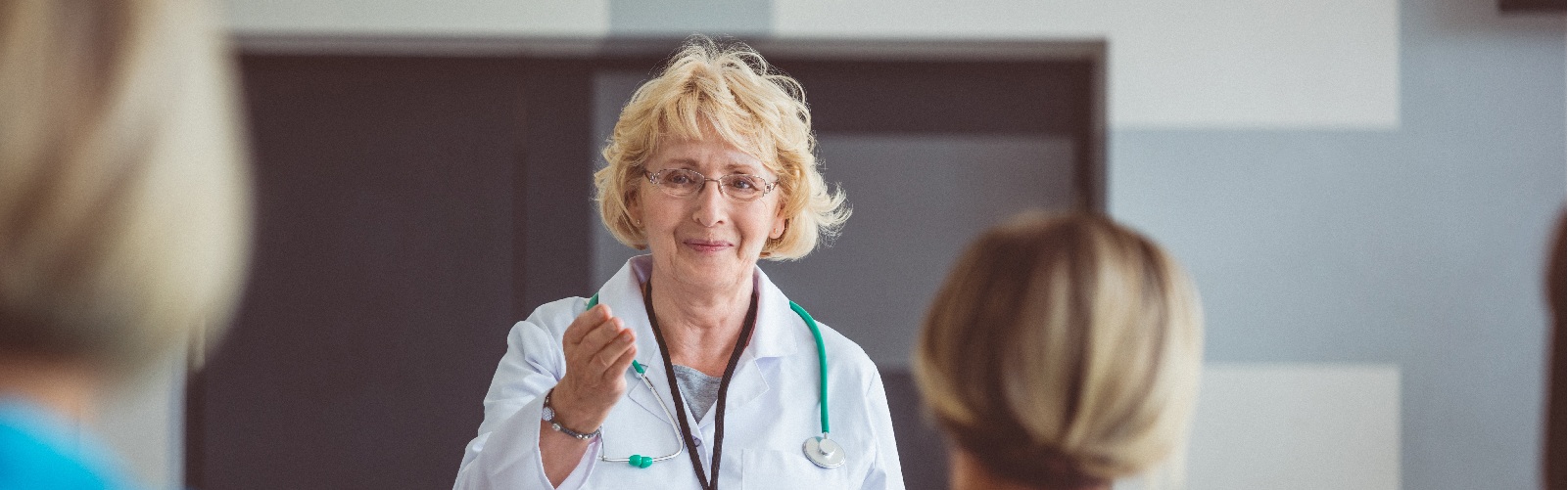 blonde female health professional in her white coat at the front of a room teaching