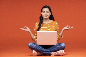 young woman with long dark hair sitting cross legged with a laptop on her legs meditating