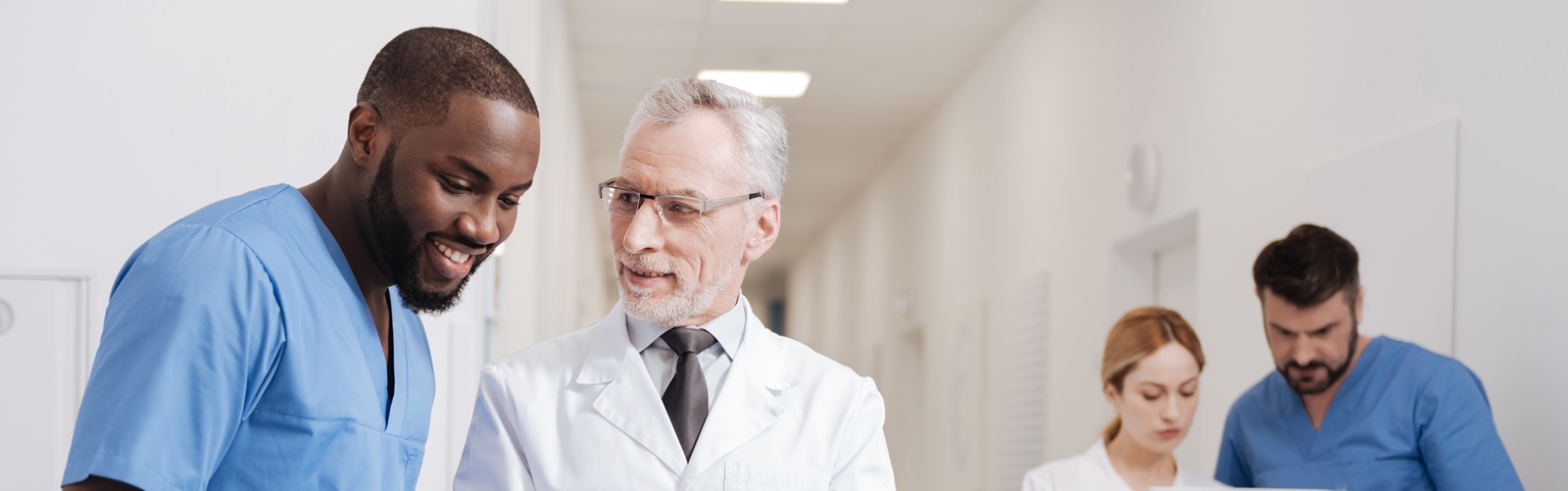 two healthcare workers discussing something in the forefront while 2 other healthcare workers look at a laptop in the background of the hallway
