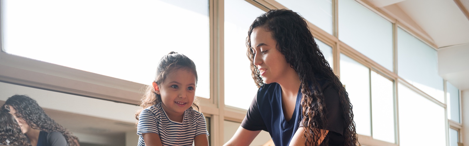 young woman in blue scrubs with long brown curly hair helping a young child balance on a pink balance ball