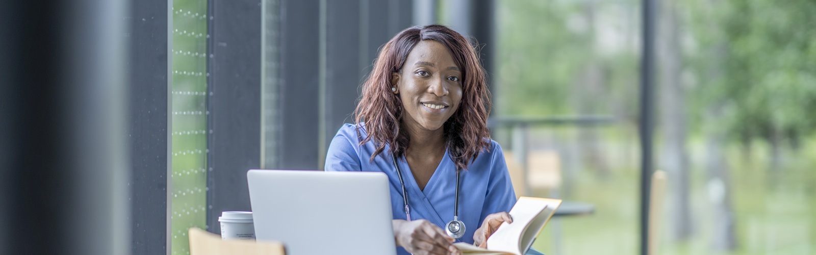 african american woman with long brown hair wearing blue scrubs is sitting at a table with an open book and a laptop