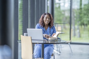 african american woman with long brown hair wearing blue scrubs is sitting at a table with an open book and a laptop