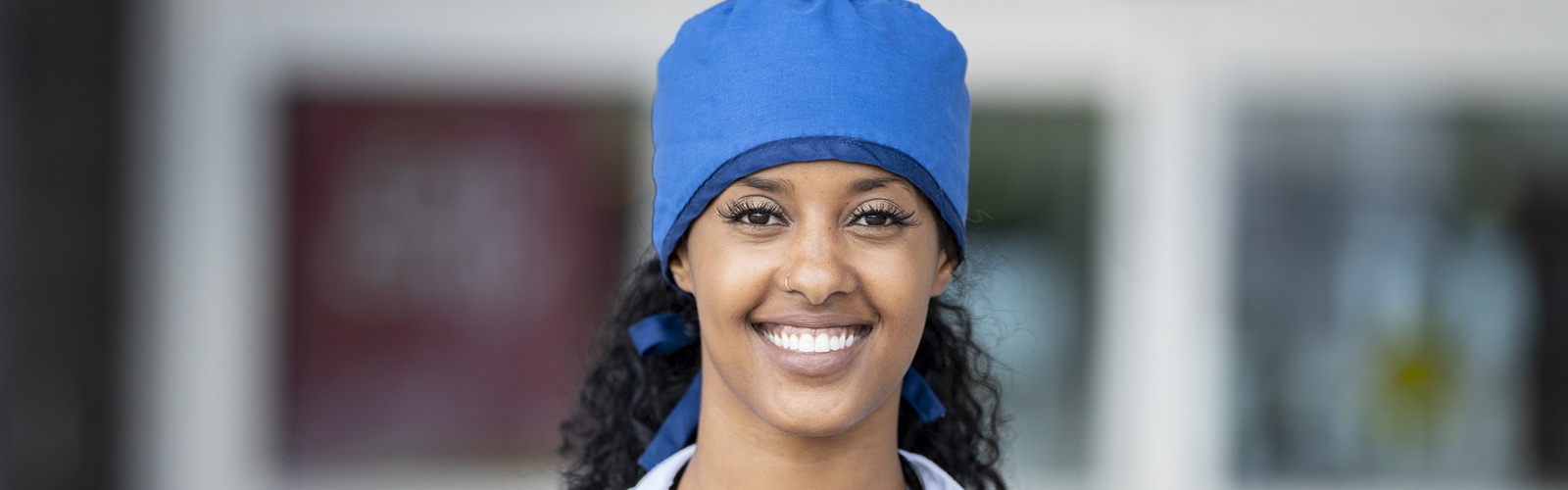 african american woman in scrubs and a white lab coat with a blue cap standing in a hallway