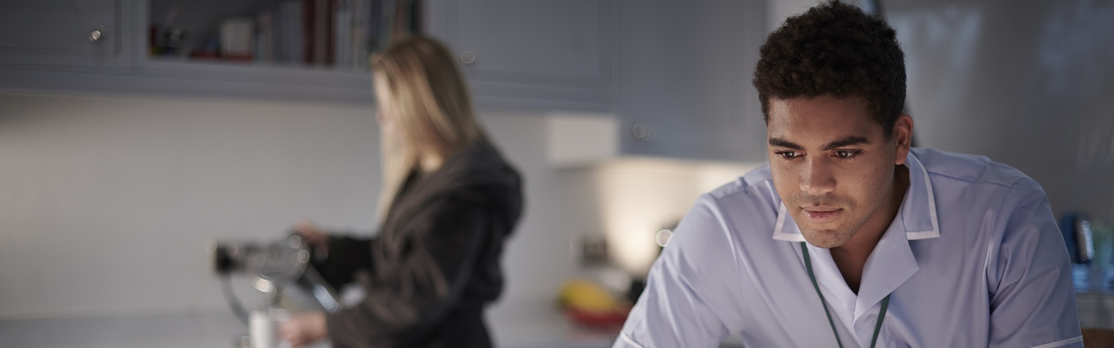 man in a blue shirt looking at a laptop with a blonde woman in a bathrobe in the background pouring coffee