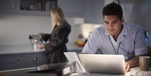 man in a blue shirt looking at a laptop with a blonde woman in a bathrobe in the background pouring coffee