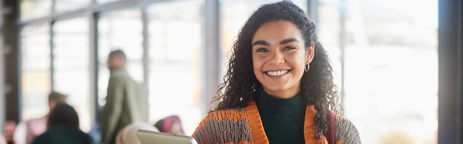 young woman with long brown curly hair and chunky orange sweater is holding a laptop standing in a classroom