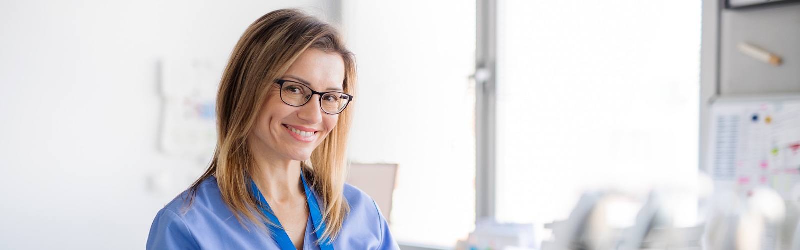 woman in blue scrubs and glasses writing on a clipboard in a hospital room