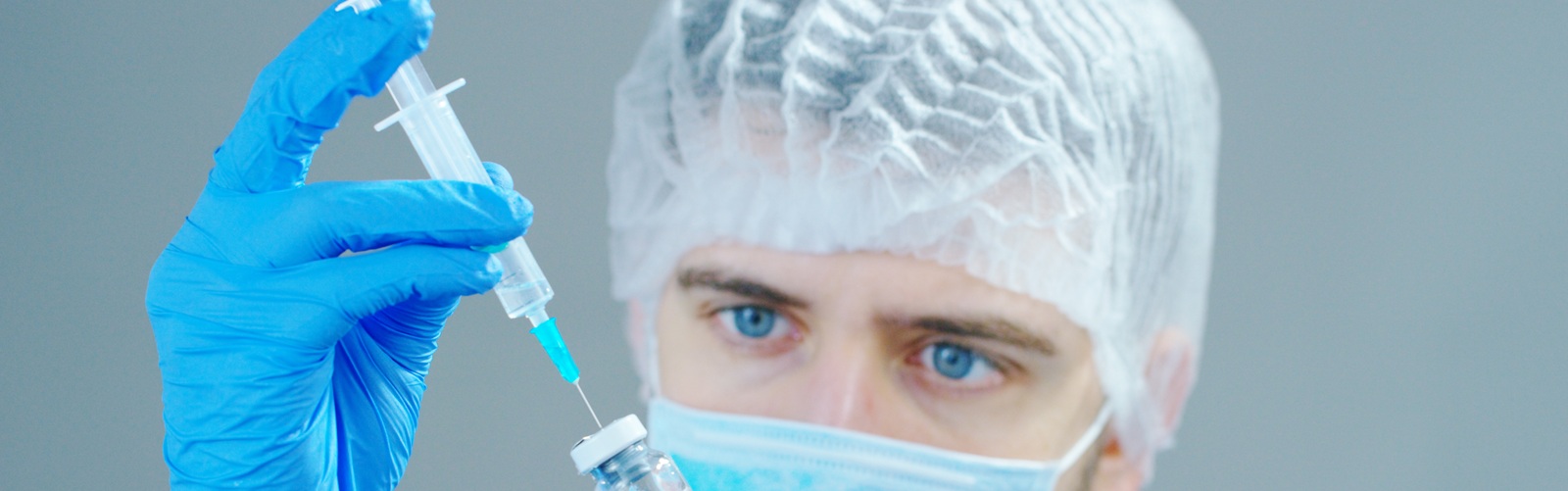 man in surgical scrubs, cap, and gloves holding a syringe and a bottle