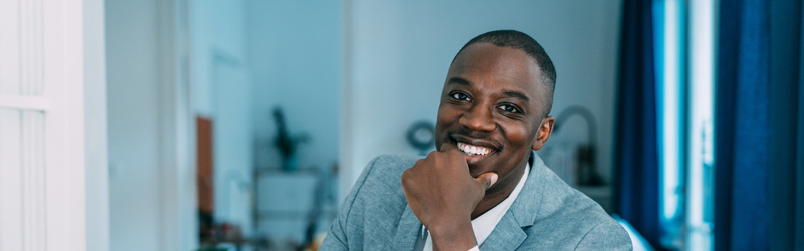 african american man in a grey jacket and white shirt smiling sitting at a desk with a laptop