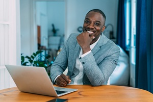 african american man in a grey jacket and white shirt smiling sitting at a desk with a laptop
