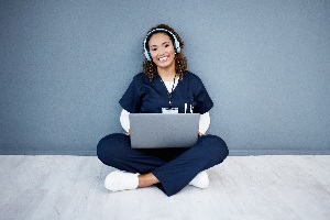 young lady with brown hair in blue scrubs sitting on the floor with a laptop in her lap