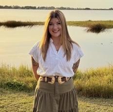 Betty Sangle headshot in a white blouse and beige skirt with a marshy lake in the background