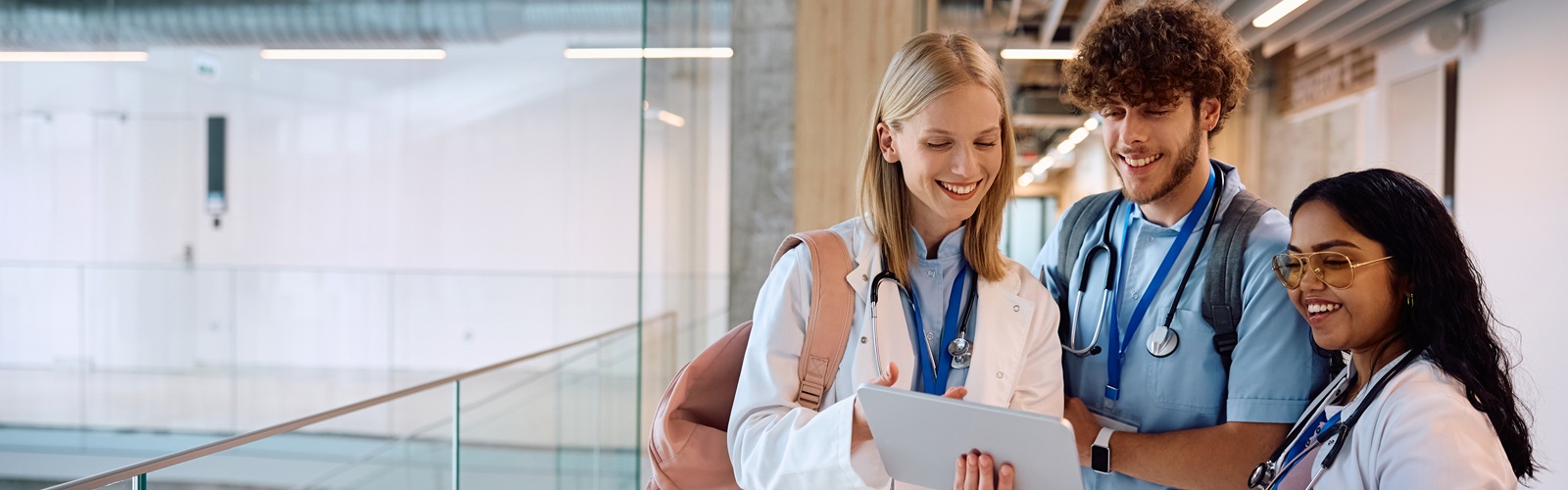 two young women, one blond and one dark haired, and one young man in a hallway wearing blue scrubs discussing paperwork
