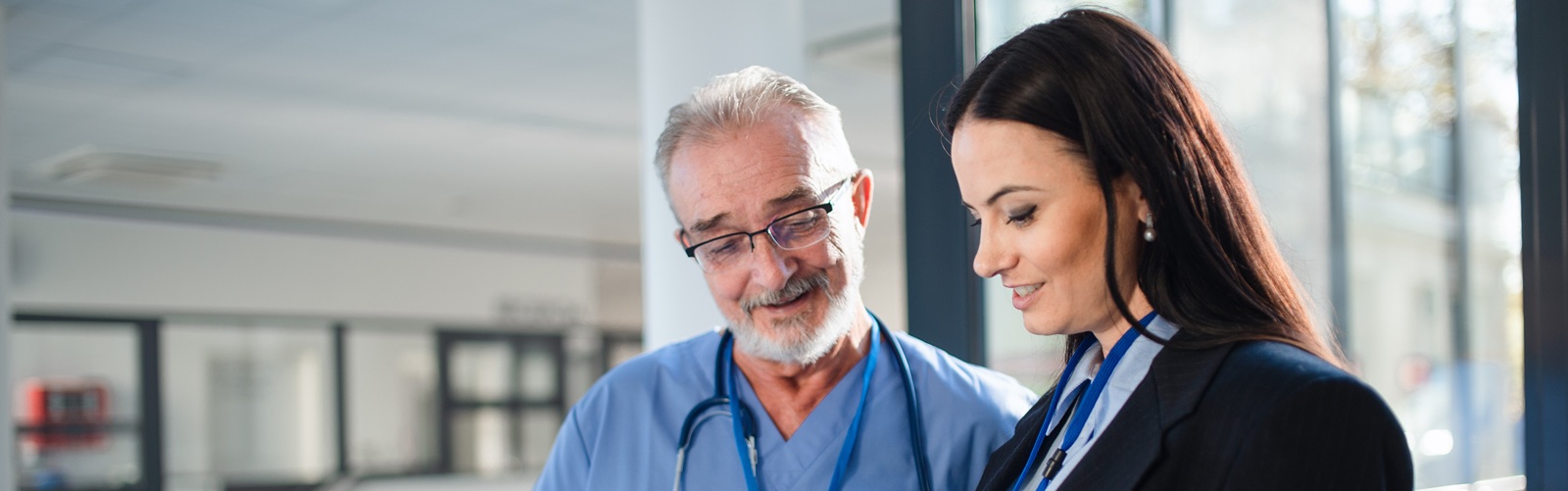 older white man in blue scrubs and younger woman with long brown hair discussing something on a tablet standing in a healthcare facility