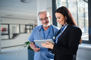 older white man in blue scrubs and younger woman with long brown hair discussing something on a tablet standing in a healthcare facility