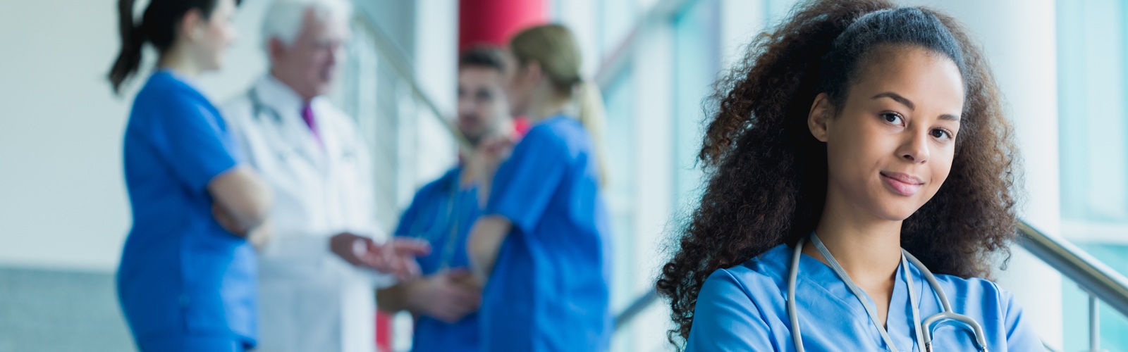 a nurse in blue scrubs standing in a hallway in front of a group of other healthcare workers in scrubs who are talking to each other