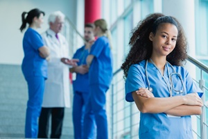 a nurse in blue scrubs standing in a hallway in front of a group of other healthcare workers in scrubs who are talking to each other