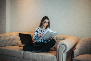 brown haired woman sitting crossed legged on a couch in a blouse and slacks with a laptop on her lap looking at papers