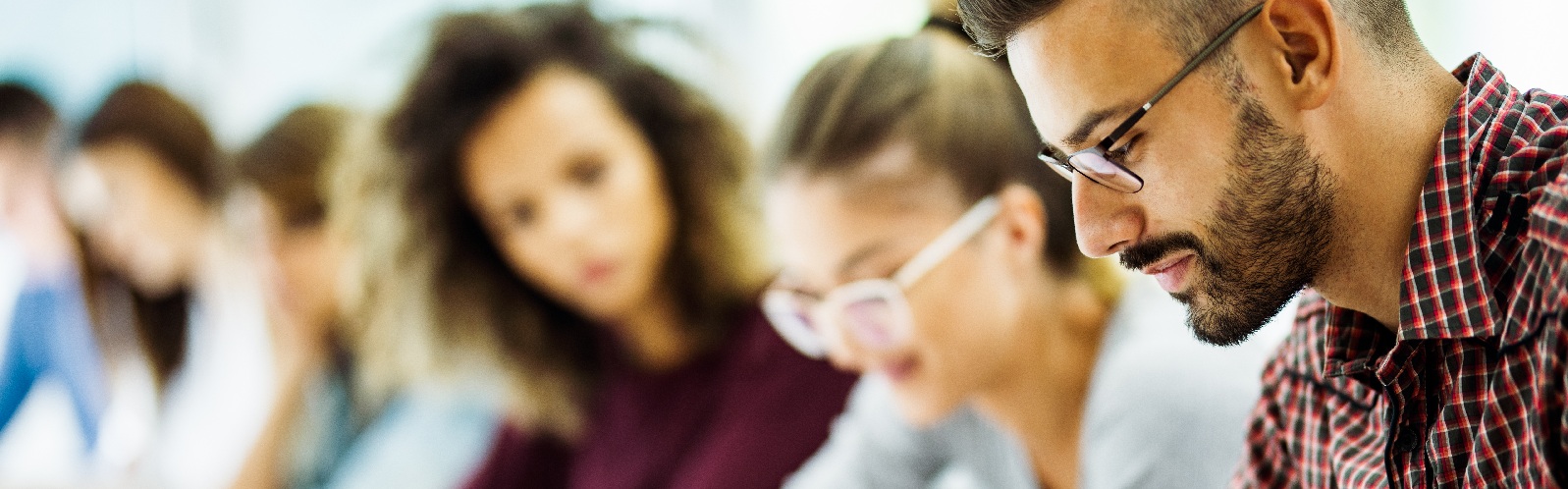 man with a slight beard and checkered button down shirt reading a book with additional classmates in the background studying
