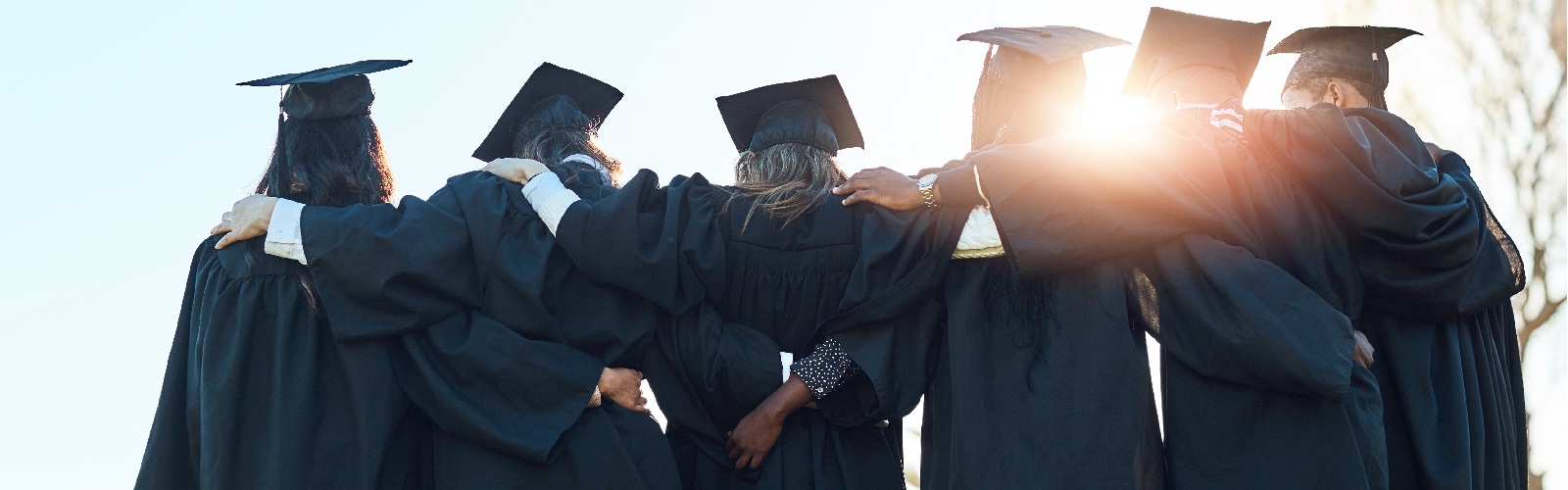 6 recent graduates holding onto each other all while still wearing their caps and gowns with the sun rays shining