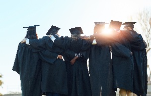 6 recent graduates holding onto each other all while still wearing their caps and gowns with the sun rays shining