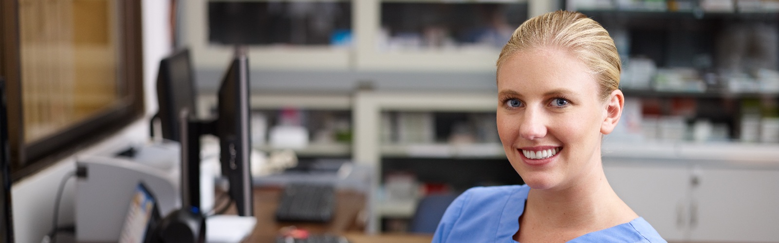 blonde woman in scrubs sitting in front of a computer in a hospital office