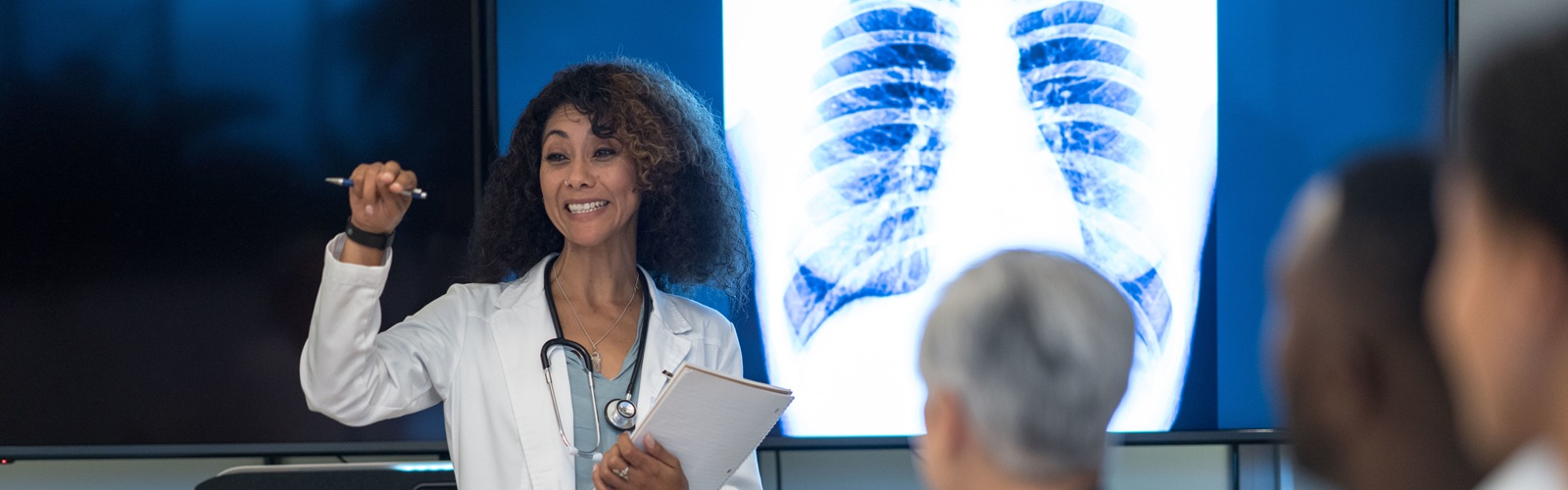 african american woman in a white doctor coat presenting to a room of other healthcare workers with an xray of a rib cage behind her