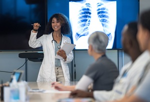 African American woman in a white doctor coat presenting to a room of other healthcare workers with an X-ray of a rib cage behind her