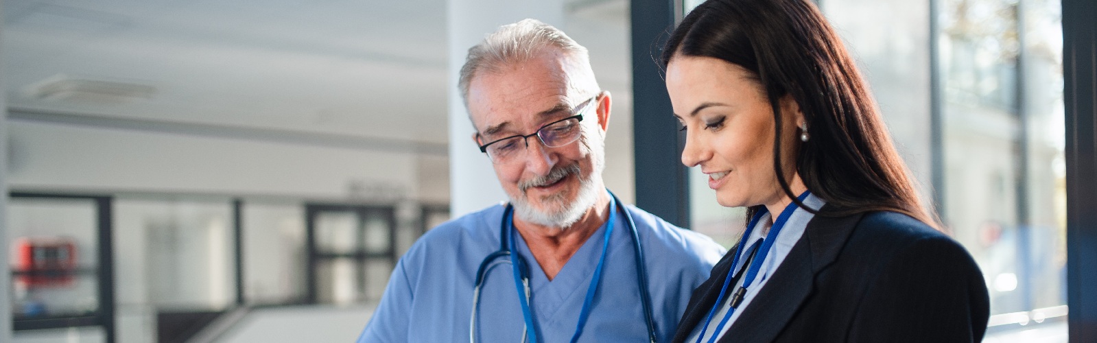 man wearing scrubs and a stethoscope and woman in a suit having a discussion in a hallway