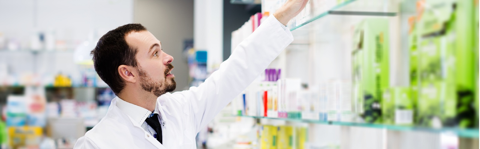 male pharmacist in a white coat reaching for something on a high shelf of a pharmacy