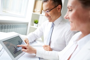 a man and a woman both in white shirts sitting together at a desk looking at a tablet screen together