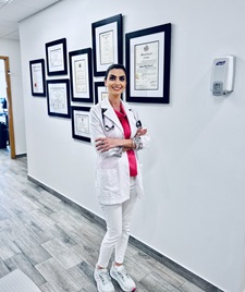 Roya Asghari standing in a hallway with multiple diplomas on the wall behind her, wearing a white coat and pink blouse