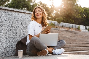 young brunette lady sitting cross legged near stairs with a laptop in her lap smiling