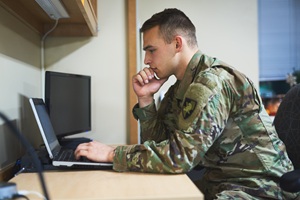 Male military member in uniform sitting at a desk looking at a laptop screen
