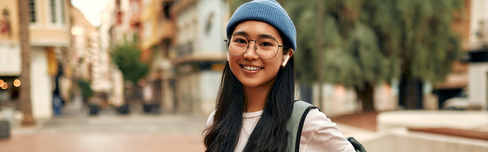 young lady with long dark hair carrying a backpack and wearing air pods holding a laptop standing in front of a building