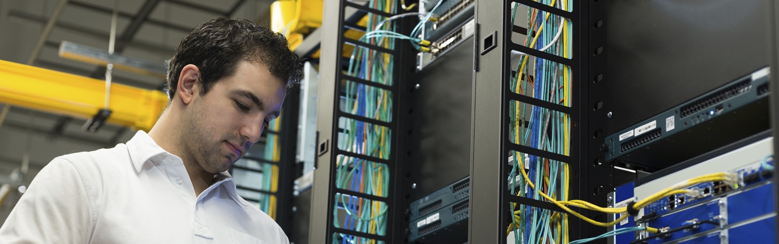 dark haired man in a white polo looking at a tablet in a server room