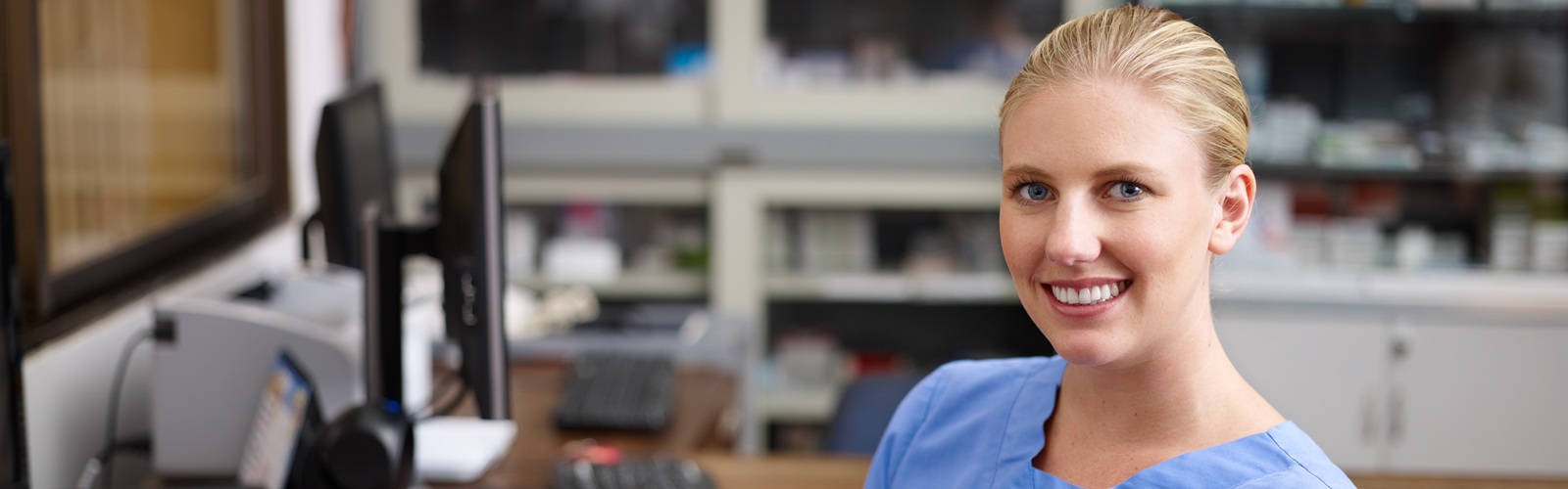 young blonde lady sitting at a desk in blue scrubs with a desktop computer