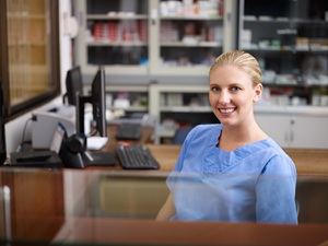 young blonde lady sitting at a desk in blue scrubs with a desktop computer