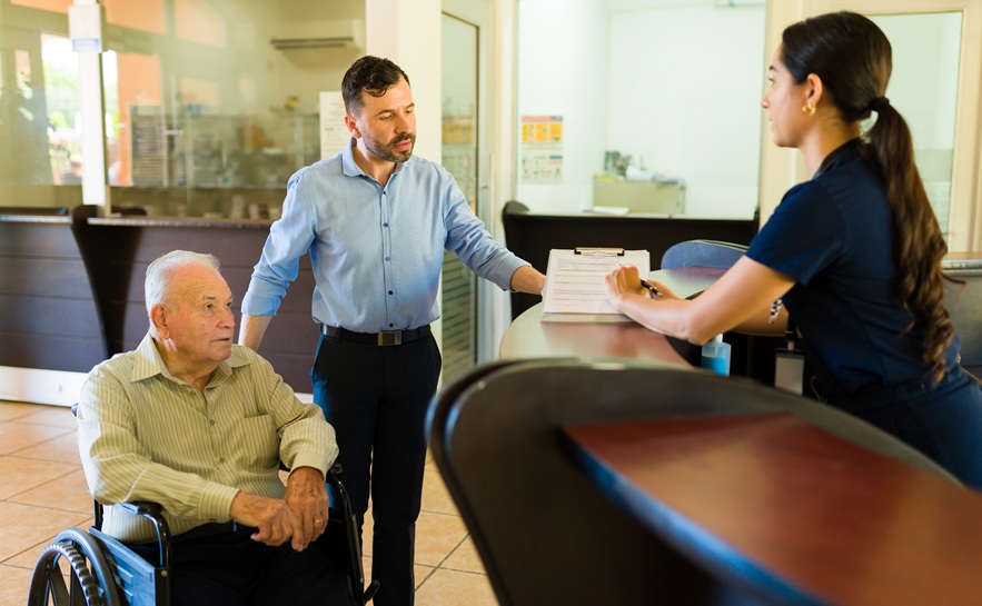female hospital receptionist standing behind a desk showing 2 men, one older in a wheelchair, paperwork on a clipboard