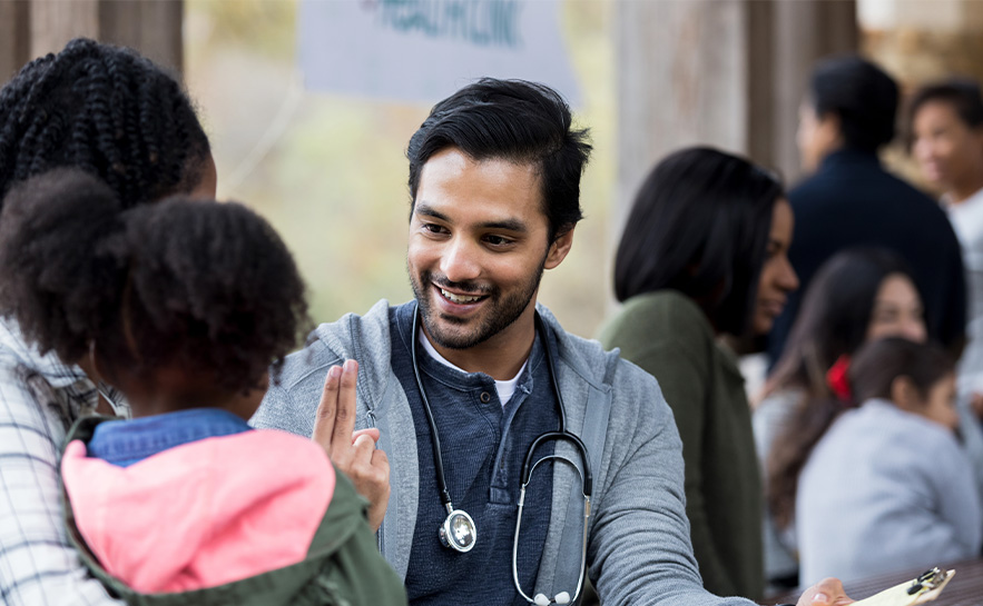 darked haired man with a short beard in a grey sweater and stethoscope holding a clipboard is speaking with a young girl and her mother