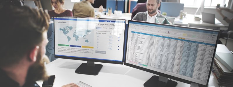 Male accountant in front of 2 computer monitors reviewing spreadsheet data